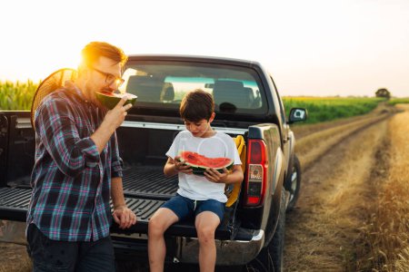 Téléchargez les photos : Father and son are enjoying a watermelon in the fields at sunset - en image libre de droit
