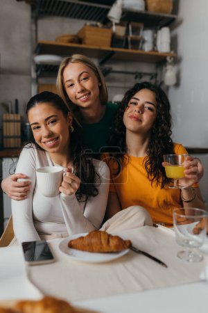 Téléchargez les photos : Vue de face de trois amis proches assis dans la cuisine et regardant la caméra - en image libre de droit