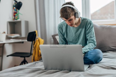 Téléchargez les photos : Adolescent garçon est assis sur l 'lit avec son ordinateur portable - en image libre de droit