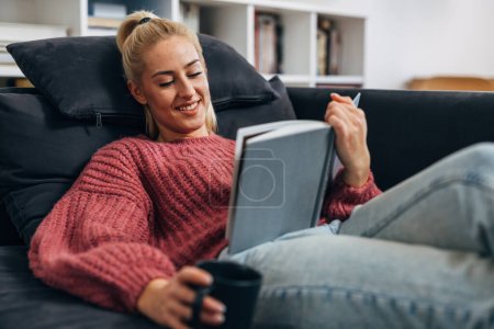 Photo for A blond Caucasian woman relaxes relaxes on the sofa with a book - Royalty Free Image