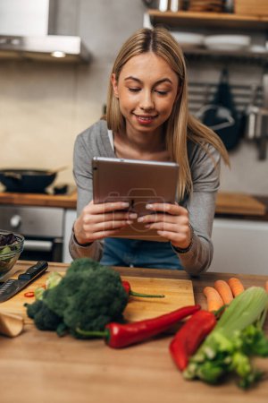 Téléchargez les photos : Vue de face d'une femme blonde dans la cuisine lisant la recette sur tablette - en image libre de droit