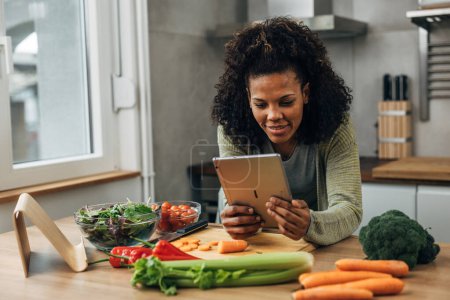 Téléchargez les photos : Heureuse femme métissée se penche sur la table de cuisine et regarde tablette - en image libre de droit