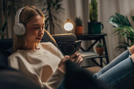 Photo for A young caucasian woman listens to music at home and looks at her phone - Royalty Free Image