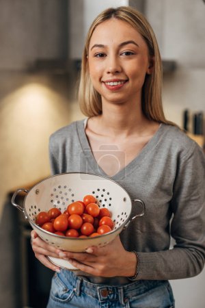 Foto de Una linda mujer caucásica sostiene un colador con tomates cherry y mira a la cámara - Imagen libre de derechos