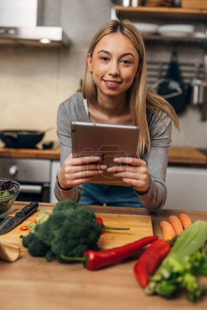 Photo for Young Caucasian woman is in the kitchen with tablet, looking at the camera - Royalty Free Image