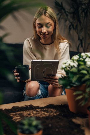 Photo for A young woman sits surrounded by potted plants and uses a digital tablet - Royalty Free Image