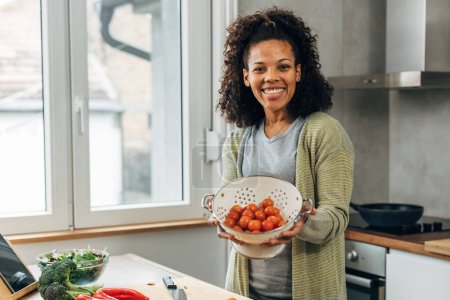 Photo for A beautiful happy middle aged woman is standing in the kitchen and holding a strainer with cherry tomatoes in it - Royalty Free Image