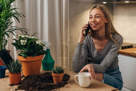 Photo for A beautiful young woman is potting her houseplants and having a pleasant conversation over the phone - Royalty Free Image