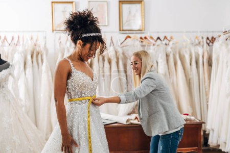 Foto de Un sastre caucásico está midiendo el tamaño de la cintura en una hermosa novia multirracial en un vestido de novia - Imagen libre de derechos