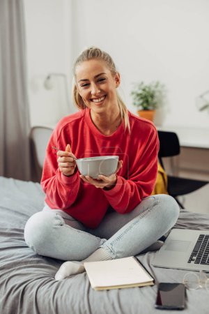 Photo for Front view of a cute college student sitting in a bed and eating oatmeal - Royalty Free Image