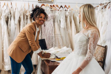 Téléchargez les photos : Femme multiraciale travaille dans un salon de mariée. Elle aide la mariée à choisir une robe de mariée. - en image libre de droit
