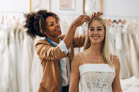 Photo for A beautiful Multiracial woman works n a bridal salon and helps a bride choose decoration that goes with her wedding dress. - Royalty Free Image