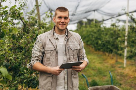 Téléchargez les photos : Heureux agriculteur caucasien surveille la ferme fruitière avec une tablette. - en image libre de droit