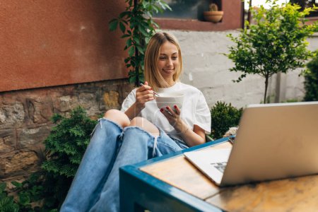Téléchargez les photos : Une belle jeune femme blonde s'assoit dans la cour et regarde un ordinateur portable - en image libre de droit