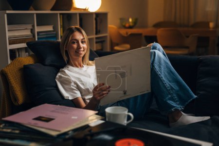 Photo for A pretty blond woman is holding a vinyl record and relaxing on the sofa with music. - Royalty Free Image