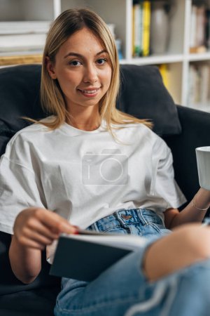 Téléchargez les photos : Une jeune femme blonde est assise sur le canapé et lit un livre. - en image libre de droit