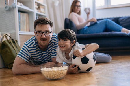 Photo for Father and son are watching a soccer game on the floor while mother sits on the sofa and looks at them. - Royalty Free Image