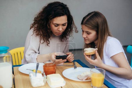 Téléchargez les photos : Une femme et une adolescente ont des sandwichs pour le petit déjeuner et regardent le téléphone mobile. - en image libre de droit