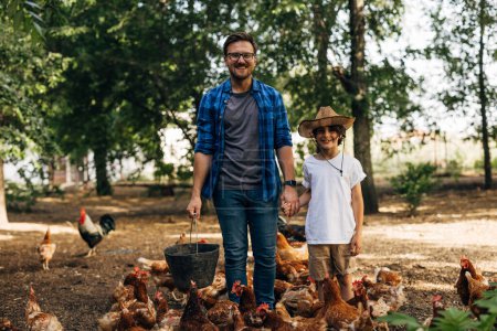 Photo for Front view of a father and son standing in their farm surrounded by hungry chickens waiting for food. - Royalty Free Image