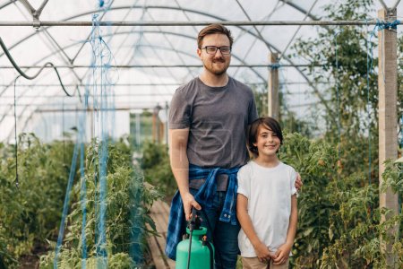 Foto de Padre e hijo de pie en la plantación de vegetales en el invernadero y mirando a la cámara. - Imagen libre de derechos