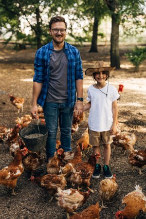 Foto de Padre e hijo tomados de la mano rodeados de gallinas afuera. - Imagen libre de derechos