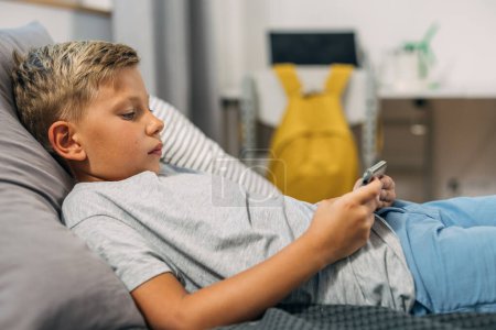 Téléchargez les photos : Vue latérale d'un jeune garçon caucasien jouant à des jeux vidéo au lit. - en image libre de droit