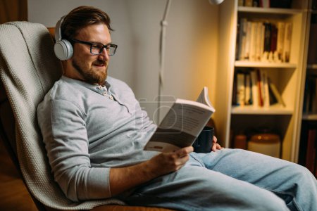 Téléchargez les photos : Un homme avec des écouteurs lisant un livre à la maison - en image libre de droit