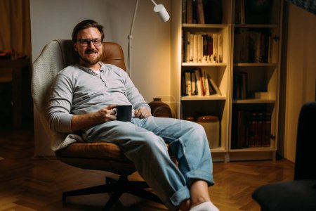 Photo for Portrait of a happy man sitting in the leather chair at home and drinking hot drink. - Royalty Free Image