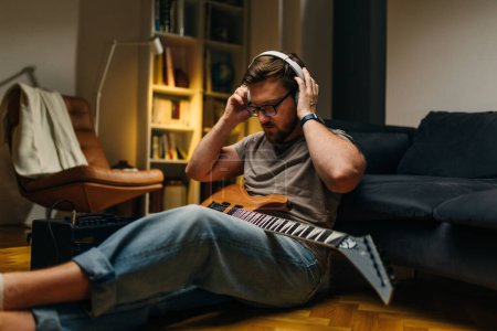 Téléchargez les photos : Musicien assis sur le sol à la maison avec une guitare électrique et mettre des écouteurs. - en image libre de droit