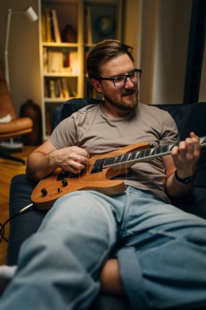 Photo for Front view of a man sitting on the sofa and playing electric guitar. - Royalty Free Image