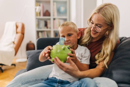 Photo for Mother and son sitting on the sofa and putting cash in a piggy bank. - Royalty Free Image
