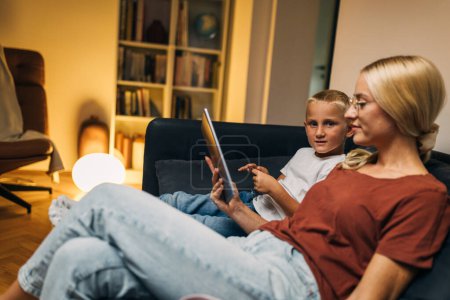 Photo for A woman is sitting on the couch with her son and they are using tablet. - Royalty Free Image