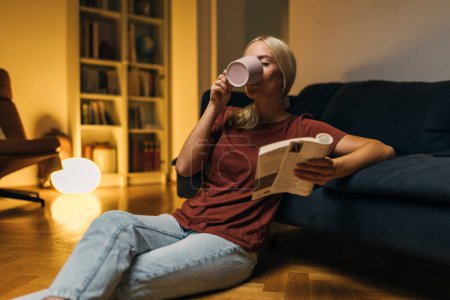 Foto de Mujer bebiendo bebida caliente y leyendo un libro en el suelo de su salón. - Imagen libre de derechos