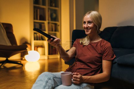 Photo for Happy woman sitting on the floor of her living room and watching television at home. - Royalty Free Image