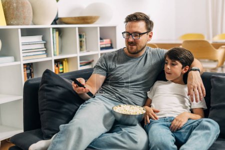 Photo for Father and son are sitting together on the sofa and watching television. - Royalty Free Image