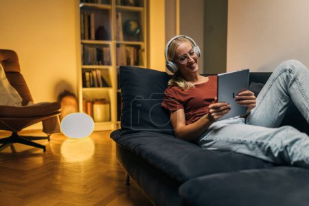 Téléchargez les photos : Belle jeune femme assise sur un canapé et utilisant une tablette numérique le soir. - en image libre de droit