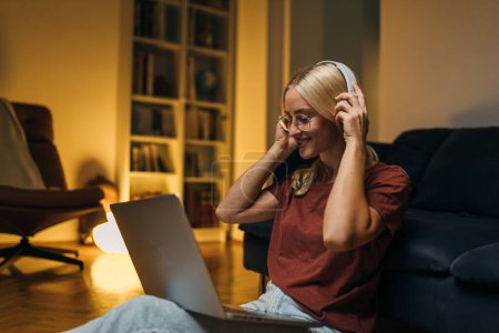 Foto de Hermosa mujer caucásica sentada en el suelo con una computadora portátil en su regazo y poniendo auriculares. - Imagen libre de derechos