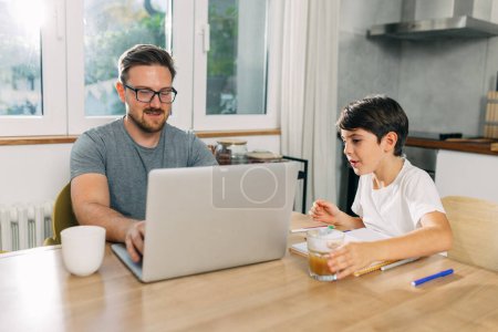 Téléchargez les photos : Père est assis dans la cuisine et travaille sur un ordinateur portable et son fils dessine à côté de lui. - en image libre de droit