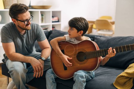 Photo for Cute Caucasian boy is learning how to play the guitar with his father. - Royalty Free Image