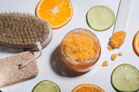 An orange scrub in a glass jar on a white table with pieces of citrus fruits. Nearby is a brush made of natural bristles for body peeling, a pumice stone for feet.
