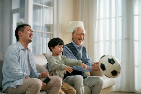 Photo for Asian son father grandfather sitting on couch at home getting angry and frustrated while watching live broadcasting of football match on TV together - Royalty Free Image