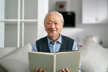 Foto de Feliz asiático viejo hombre sentado en sofá en casa leyendo un libro mirando a la cámara sonriendo - Imagen libre de derechos