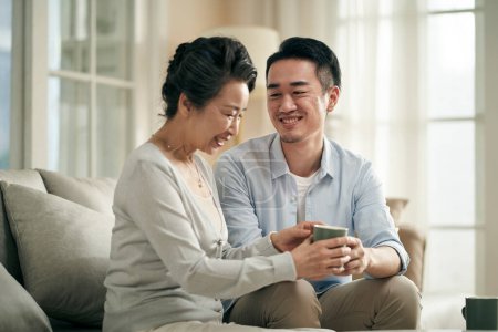 Photo for Asian adult son sitting on couch at home chatting with senior mother happy and smiling - Royalty Free Image