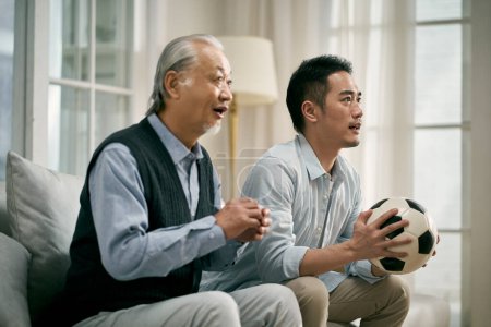 Téléchargez les photos : Père asiatique senior et fils adulte assis sur le canapé à la maison regarder match de football à la télévision ensemble - en image libre de droit
