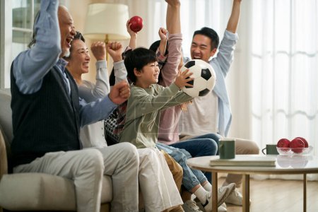 Foto de Tres generación asiático familia sentado en sofá en casa celebrando gol y victoria mientras viendo en vivo difusión de fútbol partido - Imagen libre de derechos