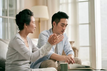 Photo for Senior asian mother and adult son sitting on couch in living room at home having a conversation - Royalty Free Image