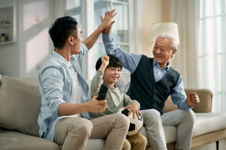 Téléchargez les photos : Asiatique fils père grand-père assis sur canapé à la maison célébrer but et la victoire tout en regardant la diffusion en direct de match de football à la télévision ensemble - en image libre de droit