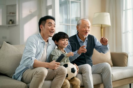Foto de Asiático hijo padre abuelo sentado en sofá en casa viendo en vivo difusión de fútbol partido en televisión juntos - Imagen libre de derechos