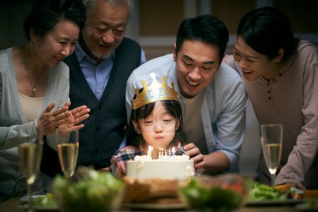 Téléchargez les photos : Petite fille asiatique soufflant des bougies tandis que la famille de trois générations fête son anniversaire à la maison - en image libre de droit