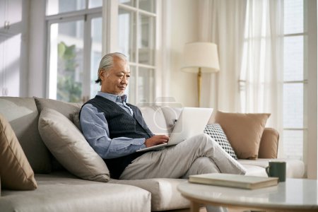 Photo for Side view of an elderly asian man sitting on couch at home using laptop computer - Royalty Free Image